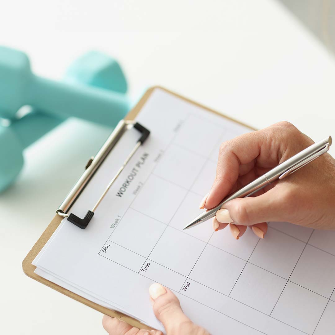 Uma pessoa preenchendo um plano de exercícios em uma prancheta com caneta prateada. Ao fundo, há um par de halteres azuis desfocados. A imagem sugere planejamento e organização de um treino intervalado para atletas de endurance.