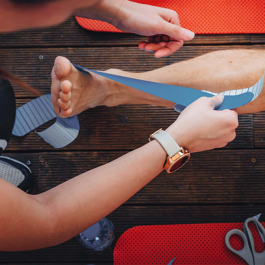 Pessoa a aplicar bandas de kinesio na perna de um atleta, possivelmente para suporte muscular ou recuperação de lesão. O ambiente parece ser um local de treino ou recuperação, com tapetes de exercício, tesoura e garrafa de água ao redor.